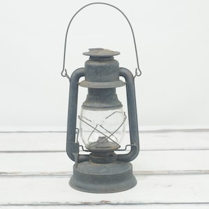 Puede incluir: Una linterna de queroseno vintage de metal gris con un globo de vidrio y un asa de alambre.