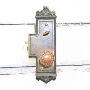May include: An antique door lock with a decorative, ornate design. The lock is made of metal with a bronze finish and features a round knob and two smaller handles. The lock is mounted on a white wooden surface.