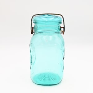 May include: A vintage aqua-colored glass jar with a wire closure. The jar has embossed detailing and a round base. The lid is the same aqua color as the jar. The wire closure is a rusty brown color. The jar is a classic design.
