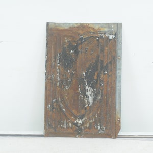 May include: A rusty metal panel with a decorative design. The panel is rectangular and has a faded white paint finish. The metal is showing signs of wear and tear, with rust and peeling paint.
