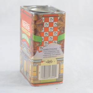 Pode incluir: Um cofre de ch&aacute; Nescaf&eacute; vintage de metal com um design vermelho, laranja e verde. O cofre apresenta um edif&iacute;cio com uma torre de rel&oacute;gio e o texto "Nescafe Bank" no lado. A parte de tr&aacute;s do cofre tem instru&ccedil;&otilde;es para fazer ch&aacute; e um c&oacute;digo de barras.