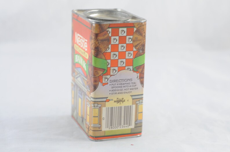 Pode incluir: Um cofre de ch&aacute; Nescaf&eacute; vintage de metal com um design vermelho, laranja e verde. O cofre apresenta um edif&iacute;cio com uma torre de rel&oacute;gio e o texto "Nescafe Bank" no lado. A parte de tr&aacute;s do cofre tem instru&ccedil;&otilde;es para fazer ch&aacute; e um c&oacute;digo de barras.