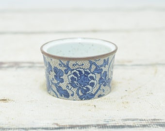 May include: A small, round ceramic bowl with a floral design. The bowl features a blue and white floral pattern on a beige background. The rim is brown. The bowl is likely used for serving or display.