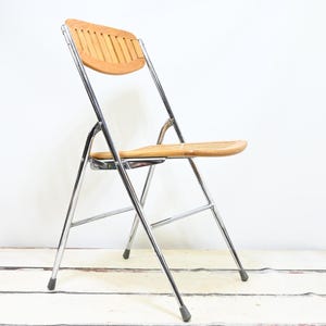 May include: A folding chair with a light brown wooden seat and backrest. The chair has a chrome-colored metal frame and black rubber feet. The chair is designed for indoor or outdoor use.