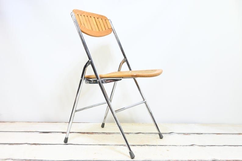 May include: A folding chair with a light brown wooden seat and backrest. The chair has a chrome-colored metal frame and black rubber feet. The chair is designed for indoor or outdoor use.