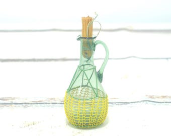 May include: A light green glass cruet with a handle and a narrow neck, featuring a yellow mesh netting covering the lower half of the body. A tan wooden stopper is inserted into the top opening of the glass vessel, completing the design.