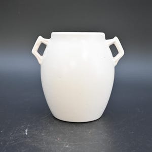 May include: A white ceramic vase with two handles. The vase has a rounded body and a wide opening. The handles are rectangular and attached to the upper part of the vase. The vase is a solid, matte white colour.