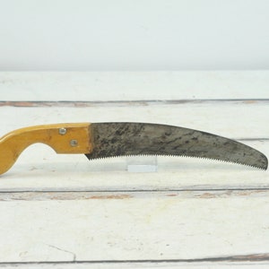 May include: A vintage hand saw with a wooden handle. The saw blade is curved and has a serrated edge. The handle is made of light brown wood and has a slight curve.