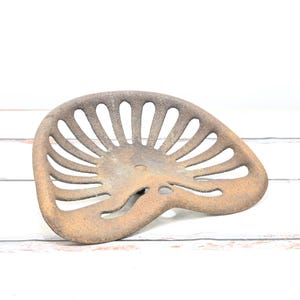 Puede incluir: Un asiento de tractor de metal oxidado con un diseño de listones. El asiento está aislado sobre un fondo de madera blanca.