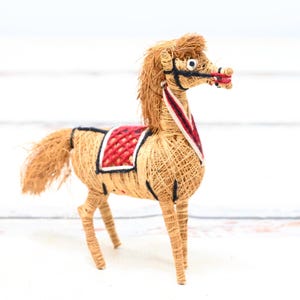 May include: A handmade horse figurine made of natural materials, with a red and black saddle. The horse is standing on a white surface.