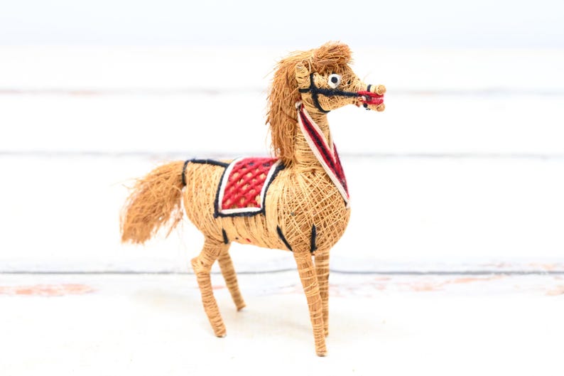 May include: A handmade horse figurine made of natural materials, with a red and black saddle. The horse is standing on a white surface.