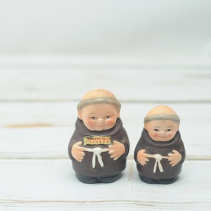May include: Two ceramic salt and pepper shakers shaped like monks. The shakers are brown and have white faces. The shaker on the left has a small sign that reads "Niagara Falls Canada".