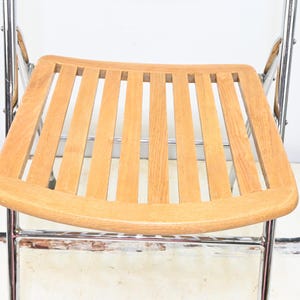 May include: A folding chair with a light brown wooden seat and backrest. The seat has a slatted design. The frame is made of shiny, silver-colored metal. The chair is set against a white background.