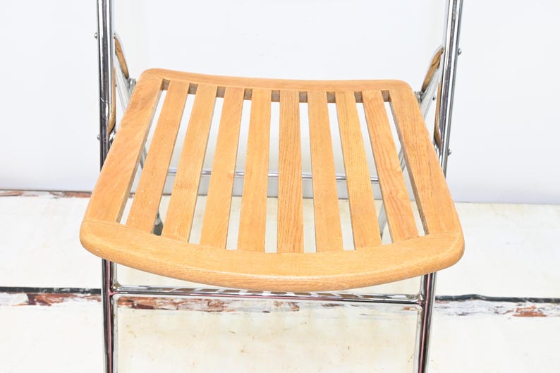 May include: A folding chair with a light brown wooden seat and backrest. The seat has a slatted design. The frame is made of shiny, silver-colored metal. The chair is set against a white background.