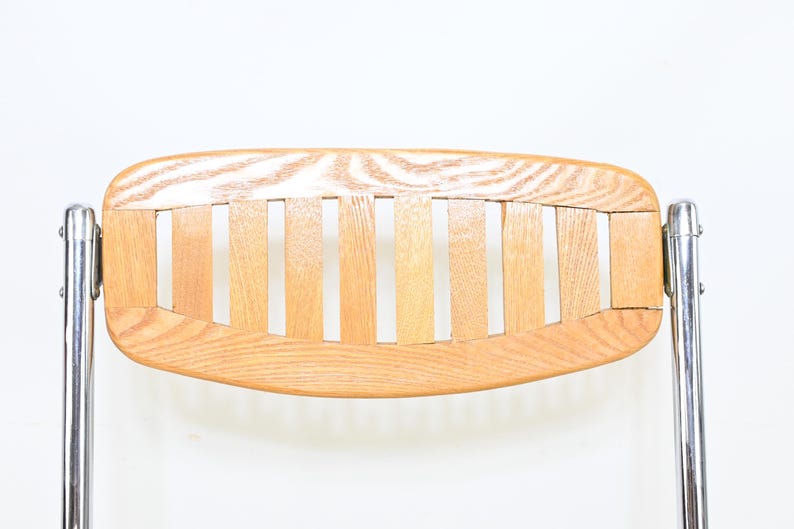 May include: Close-up of a vintage chair back. The backrest is made of light-colored wood slats and a curved wooden frame. The chair has shiny, silver metal legs. The design is minimalist and mid-century modern.