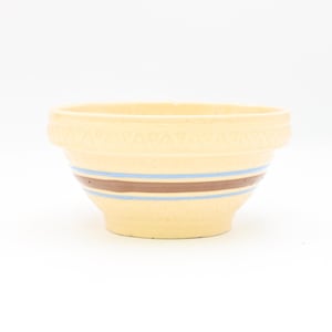 Puede incluir: Un cuenco de cerámica color crema con un borde decorativo y dos rayas horizontales en azul y marrón. El cuenco tiene forma redondeada y está sobre un fondo blanco.