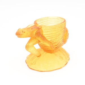 Könnte beinhalten: Ein gelber gläserner Eierbecher in Form eines Frosches. Der Frosch sitzt auf einem Blatt und hält den Eierbecher mit seinen Hinterbeinen.