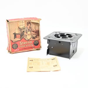 Könnte beinhalten: Ein schwarzer Sterno-Kocher aus Metall mit quadratischer Oberseite und rundem Rost, begleitet von der originalen rot-beigen Pappschachtel mit Vintage-Illustrationen und dem Text Sterno Cook Stove sowie einem beigen Papier-Anleitungsblatt.