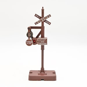 May include: A miniature brown railroad crossing signal with a base, pole, and crossbucks. The sign reads "RAIL CROSSING ROAD" and "STOP WHEN SWINGING". A red light is visible on the stop sign.