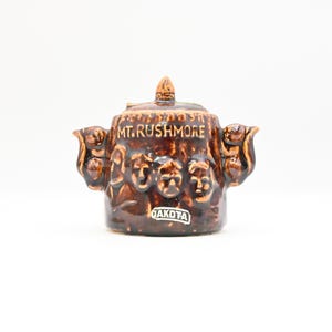 May include: A brown ceramic Mt. Rushmore souvenir teapot. The teapot features the faces of the presidents carved into the side, with the words "MT. RUSHMORE" on the top. The label "DAKOTA" is on the front.