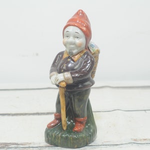 Może przedstawiać: Ceramiczna figurka krasnala w czerwonym kapeluszu i brązowych ubraniach. Trzyma laskę i ma kosz na plecach.