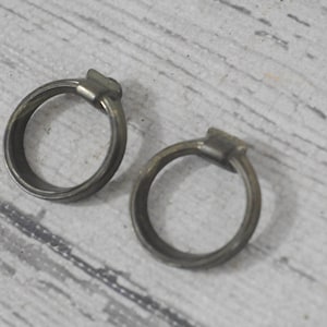 Puede incluir: Dos anillos de metal plateados con un acabado redondeado y liso. Los anillos son de tamaños ligeramente diferentes y están colocados uno al lado del otro sobre una superficie de madera clara.