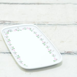 Op de afbeelding: Witte rechthoekige serveerschaal met een bloemige rand van roze rozen en groene bladeren.