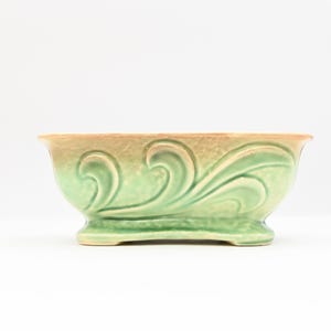 以下が含まれることがあります： 淡い緑とベージュの配色が施された、長方形のヴィンテージプランター。セラミックプランターは、側面に浮き彫りの渦巻き模様があり、小さなベースの上に置かれています。縁はベージュ色です。