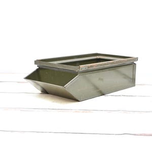 May include: An olive green metal storage bin, rectangular with a sloped front. It features a rectangular opening at the top and is set against a white wooden background. The bin is likely for storage or display purposes.