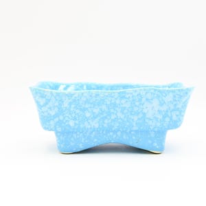 May include: A light blue ceramic planter with a speckled pattern. The rectangular planter has a curved rim and is supported by three curved feet. The planter is approximately 15 cm long, 8 cm wide, and 8 cm tall.