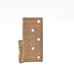 Puede incluir: Una bisagra metálica antigua y ornamentada, con una placa rectangular y un cilindro. La bisagra es de color marrón óxido con diseños florales y geométricos decorativos. Tiene varios agujeros para el montaje.