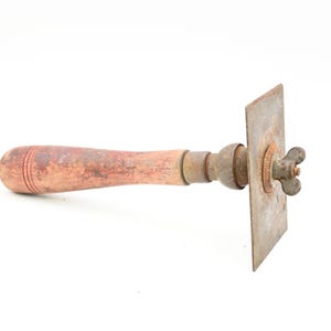 May include: An antique metal tool with a wooden handle. The handle is a reddish-brown color and has a rounded end. The metal components show signs of age and wear, with a square plate and a wingnut.