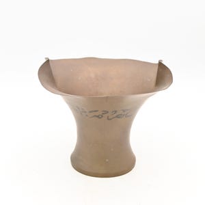 May include: A bronze-colored vase with a flared top and a rounded base. The vase has a wide, open mouth and a slightly curved rim. The body of the vase features an inscription in a stylized script. The vase is set against a white background.