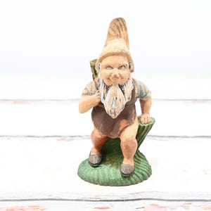 Może przedstawiać: Ceramiczna figurka krasnala z brązowym kapeluszem, białą brodą i brązowymi spodniami. Krasnal siedzi na zielonej podstawie.