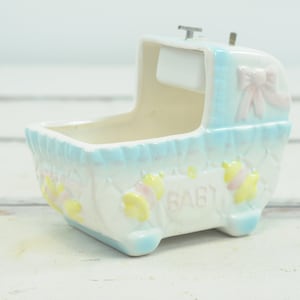 Puede incluir: Una caja de música de cerámica blanca en forma de cuna para bebé con ribete azul y blanco. La cuna tiene un lazo rosa y la palabra "BABY" escrita en la parte delantera.