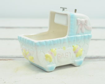 May include: A white ceramic baby crib music box with a blue and white trim. The crib has a pink bow and the word "BABY" written on the front.