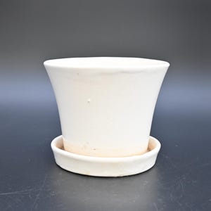 Op de afbeelding: Een witte keramische plantenpot met een gladde, licht uitlopende rand en een bijpassende ronde schotel. Het buitenoppervlak heeft een strakke, effen witte afwerking met subtiele textuurvariaties bij de onderste rand waar de pot de schotel raakt.
