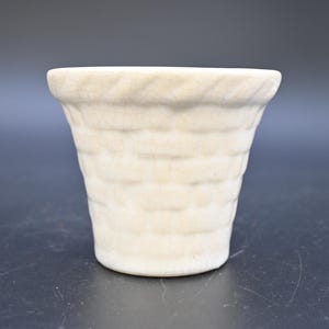 Peut inclure: Un petit pot de fleurs en céramique blanc cassé présentant un extérieur texturé avec un motif de vannerie tressée. Le récipient possède un bord évasé et un corps cylindrique qui se rétrécit légèrement vers la base, avec une finition craquelée.