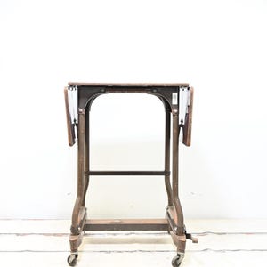 May include: An antique, brown wooden table with a metal frame and wheels. The table has a rectangular top with two drop-leaf extensions. The metal frame is dark and sturdy, and the table is on four small caster wheels.