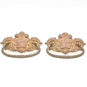 May include: Two antique bronze drawer pulls with ornate floral and scrollwork designs. Each pull features a decorative backplate with a blank rectangular space and a rope-style handle. The hardware is a warm, aged bronze color.