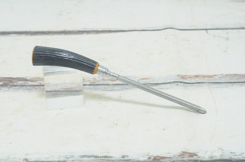 May include: A black and brown handled metal pencil sharpener with a silver metal blade. The sharpener is on a white wooden surface.