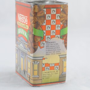 Pode incluir: Uma lata de metal vintage com um design vermelho, laranja e branco. A lata apresenta um edif&iacute;cio banc&aacute;rio com uma placa que diz "Nestl&eacute; Hot Cocoa Mix BANK". A lata &eacute; decorada com moedas e uma fita verde.