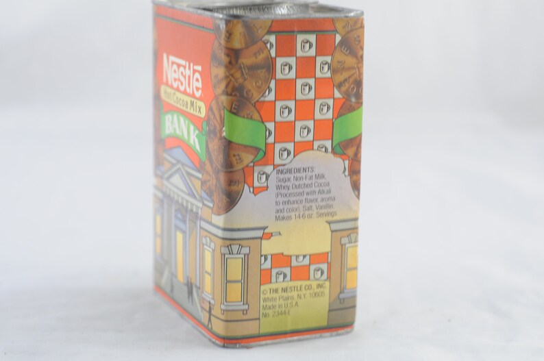 Pode incluir: Uma lata de metal vintage com um design vermelho, laranja e branco. A lata apresenta um edif&iacute;cio banc&aacute;rio com uma placa que diz "Nestl&eacute; Hot Cocoa Mix BANK". A lata &eacute; decorada com moedas e uma fita verde.