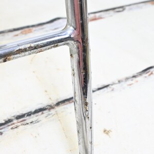 May include: Close-up of a section of a chrome chair frame. The metal has some rust and wear. The background is a white surface with black and brown markings.