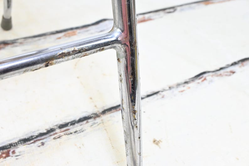 May include: Close-up of a section of a chrome chair frame. The metal has some rust and wear. The background is a white surface with black and brown markings.