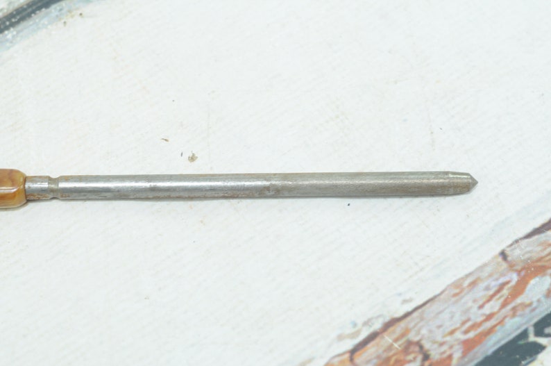 Op de afbeelding: Een close-up van een metalen gereedschap met een bruine kunststof handvat. Het gereedschap heeft een puntige punt en een lange, dunne schacht.