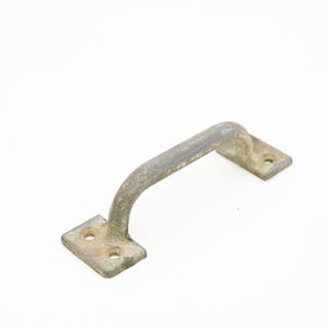 May include: A weathered, antique-style metal handle with a curved grip and two rectangular mounting plates. The handle is a muted, aged brass color, showing signs of wear and use. Each plate has two screw holes.