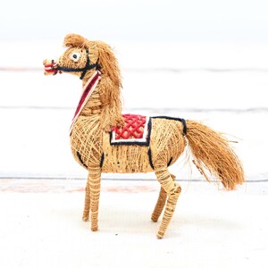 May include: A handmade horse figurine made of natural materials like straw and twine. The horse is decorated with a red and white saddle and has a brown mane and tail.