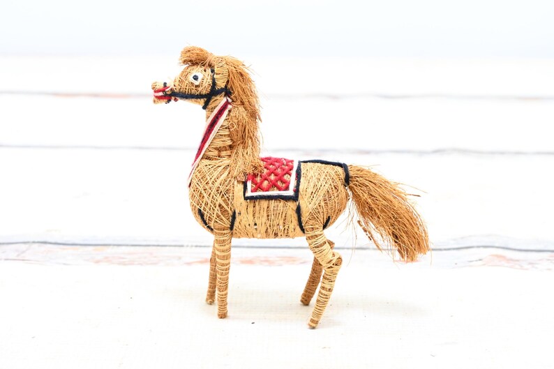May include: A handmade horse figurine made of natural materials like straw and twine. The horse is decorated with a red and white saddle and has a brown mane and tail.