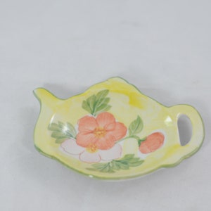 Puede incluir: Un plato de cerámica amarillo en forma de tetera con un diseño floral. El plato presenta una flor rosa y naranja con hojas verdes y un fondo amarillo.